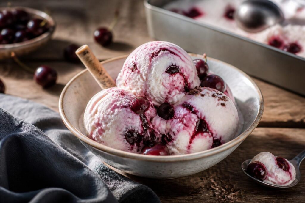 Kirsch-Quark-Eis ohne Eismaschine in einer cremefarbenen Gefrierbox, mit glänzenden Kirschen und Minzblättern garniert, von oben fotografiert